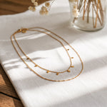 Zweireihige Halskette aus Edelstahl 18 K vergoldet mit kleinen quadratischen Anhängern, liegt auf weißem Stoff mit Holzuntergrund und Trockenblumen – Zelina Layers Halskette | Mercy’s Jewelry