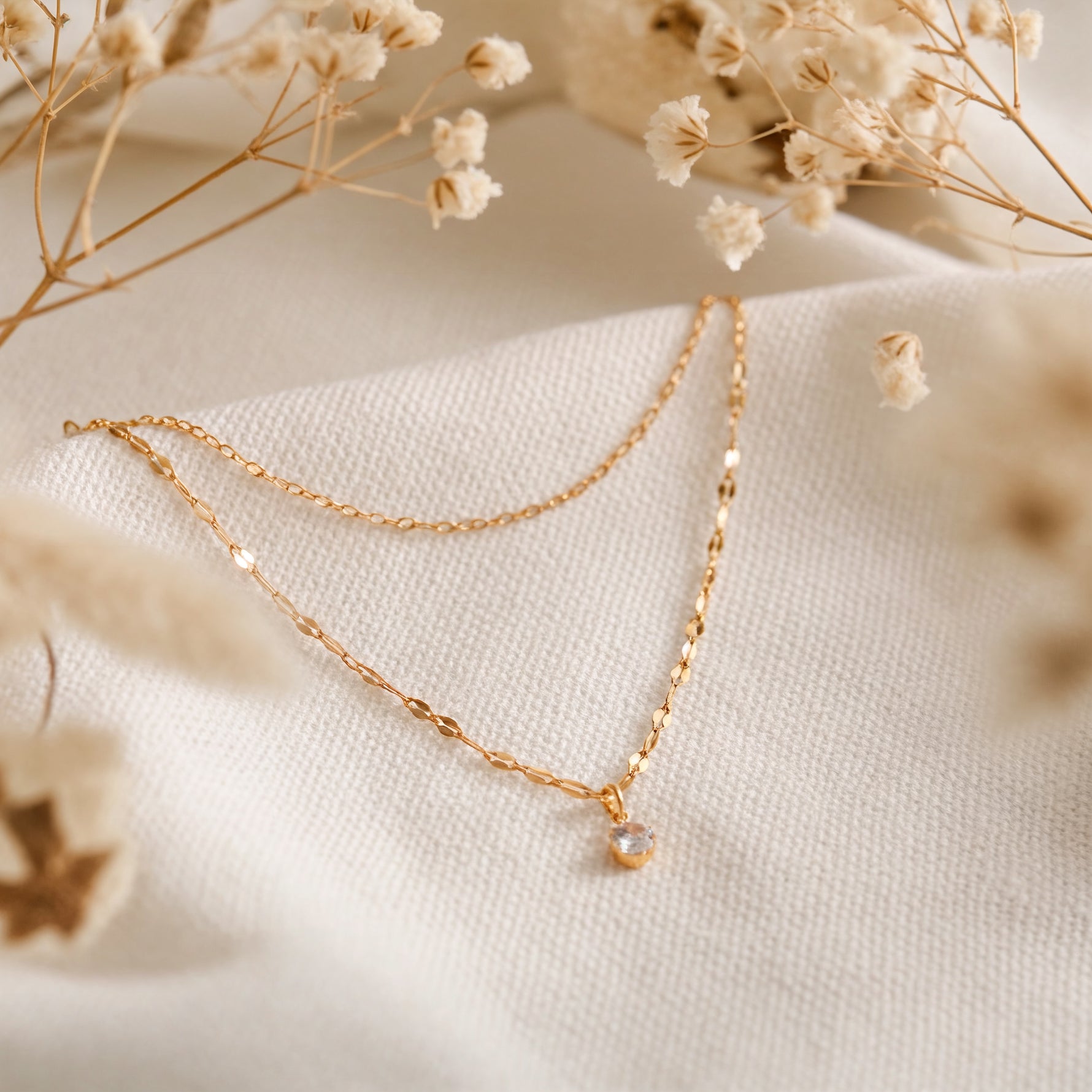 Zweireihige Halskette aus Edelstahl 18 K vergoldet mit feinem Zirkonia-Anhänger, liegt auf hellem Stoff mit Trockenblumen im Hintergrund – Lirae Layers Halskette | Mercy’s Jewelry