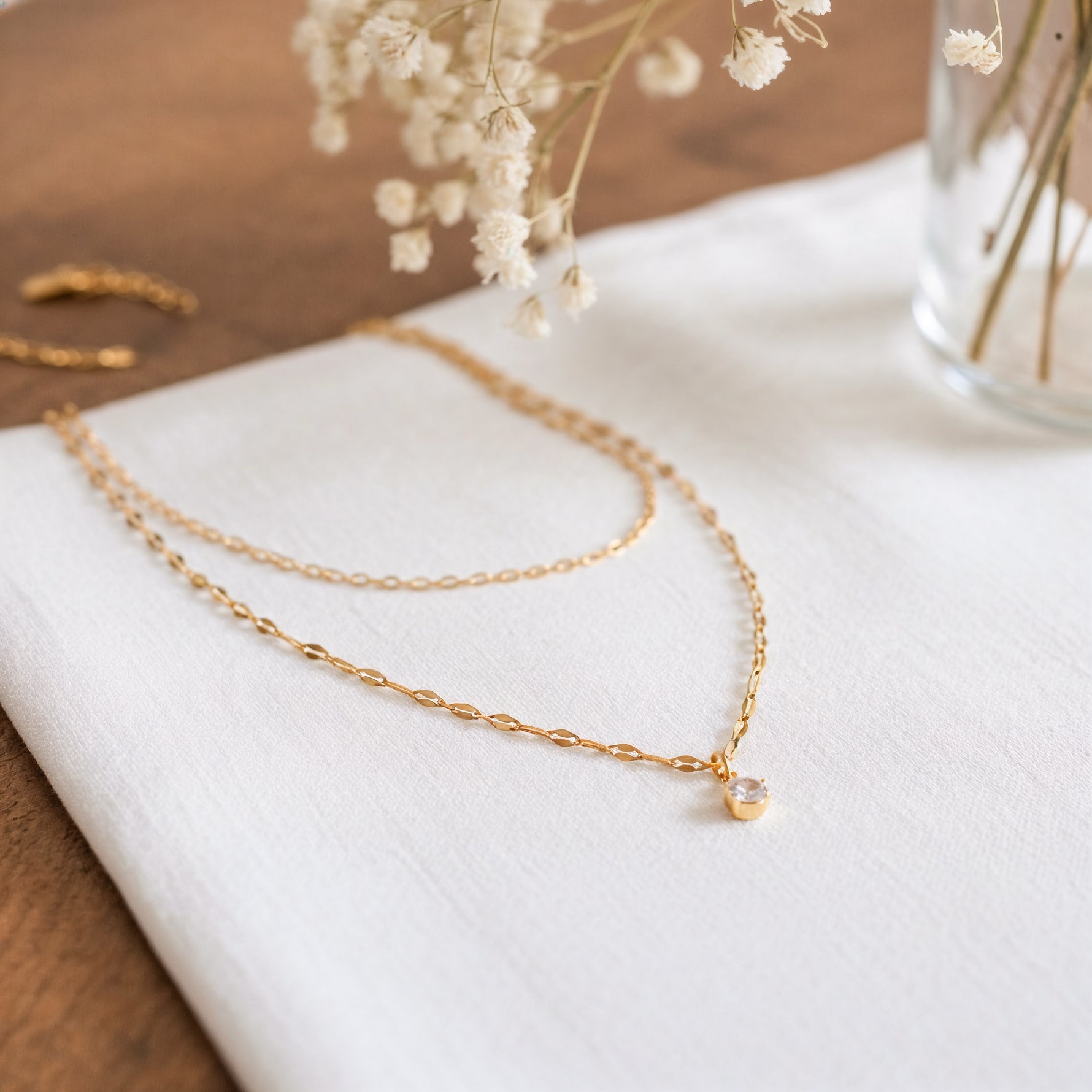 Zweireihige Halskette aus Edelstahl 18 K vergoldet mit feinem Zirkonia-Anhänger, liegt auf weißem Stoff mit Glasvase und Trockenblumen – Lirae Layers Halskette | Mercy’s Jewelry