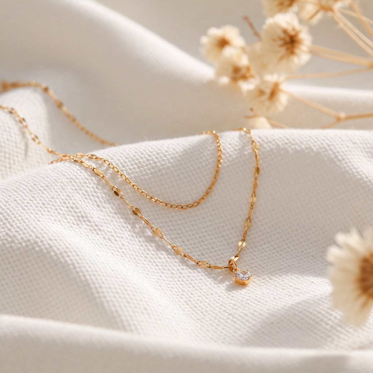 Zweireihige Halskette aus Edelstahl 18 K vergoldet mit feinem Zirkonia-Anhänger, liegt auf weißem Stoff mit Trockenblumen im Hintergrund – Lirae Layers Halskette | Mercy’s Jewelry