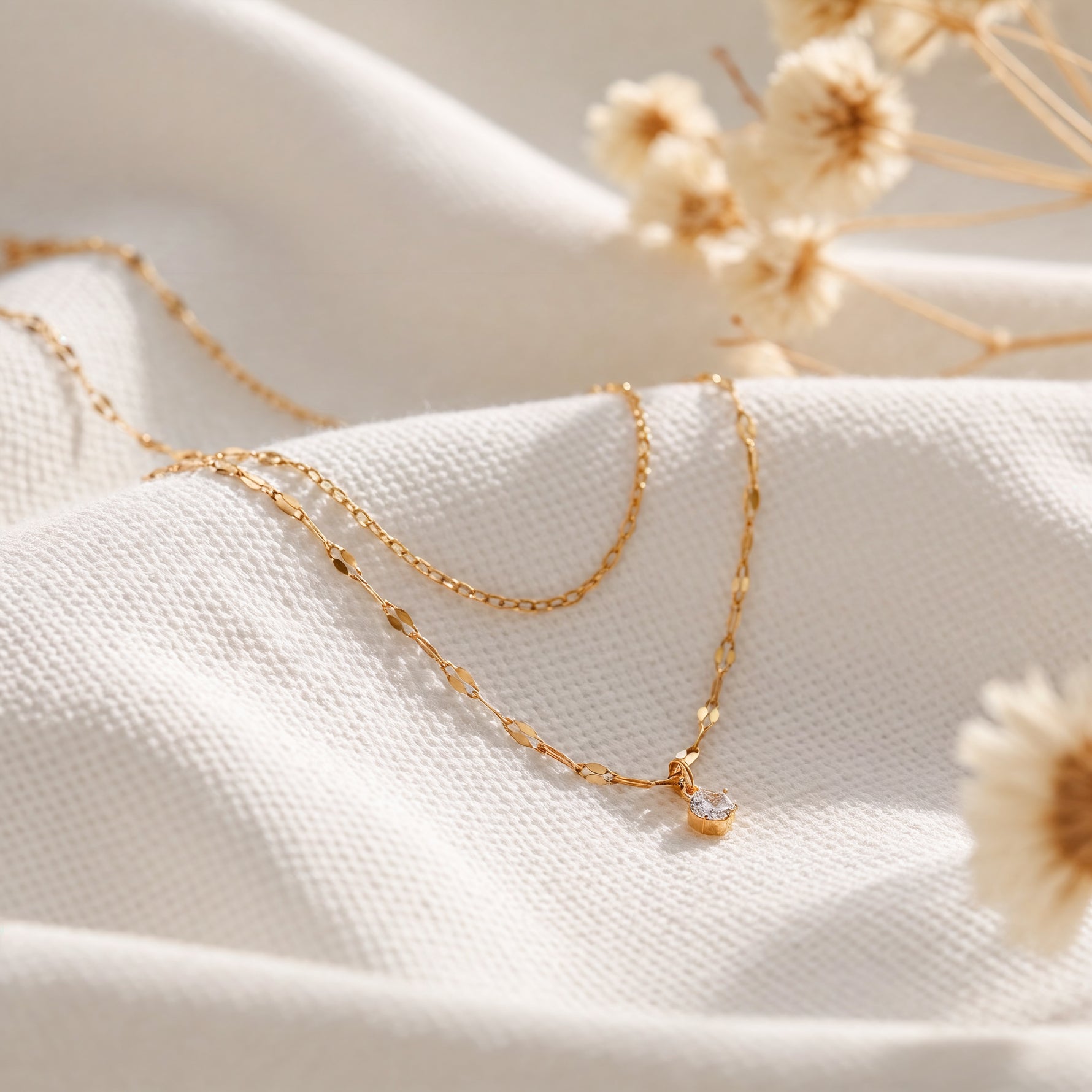 Zweireihige Halskette aus Edelstahl 18 K vergoldet mit feinem Zirkonia-Anhänger, liegt auf weißem Stoff mit Trockenblumen im Hintergrund – Lirae Layers Halskette | Mercy’s Jewelry